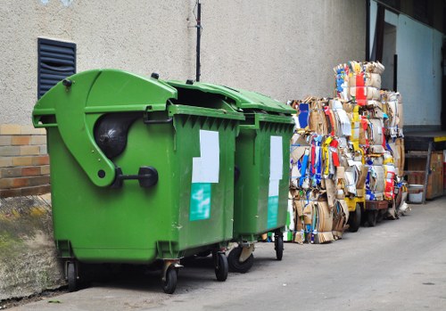 In front of a beige concrete wall, there are two large green wheelie bins with closed lids, positioned on a paved urban street. The bins are made of durable plastic with textured surfaces, one featuring a black wheel and handle. To the right of the bins, there are stacked bundles of flattened cardboard packaging, held together with plastic straps, with visible corrugated surfaces and various printed or coloured sections, indicating they contain recyclable materials. A small section of a building with a glass window and dark window frame can be seen in the background. The scene suggests waste collection in a residential or commercial area, typical of rubbish removal services in Brompton, with the bins ready for collection or disposal. The overall environment looks clean and orderly, with the rubbish organized for prompt removal by a professional service such as Commercial Waste Removal Brompton.