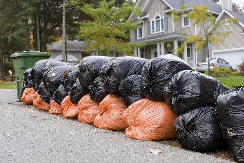 A row of black and orange rubbish bags filled with waste is stacked along the edge of a residential street, positioned on the tarmac pavement in front of a green wheelie bin. The black bags are placed on top of the orange ones, with some bags partially leaning against each other, indicating recent collection or awaiting removal. In the background, there are a few trees with fresh green leaves and a two-storey house with white and blue siding, characteristic of suburban Brompton properties. The scene is captured during daylight hours, with natural lighting casting soft shadows on the street and house. This setup illustrates typical rubbish disposal practices for household waste management, aligning with services offered by Commercial Waste Removal Brompton in the local area. The overall environment appears clean and orderly, emphasizing efficient waste collection in a residential neighbourhood, suitable for a professional rubbish removal service operating in Brompton or nearby areas with similar street scenes.