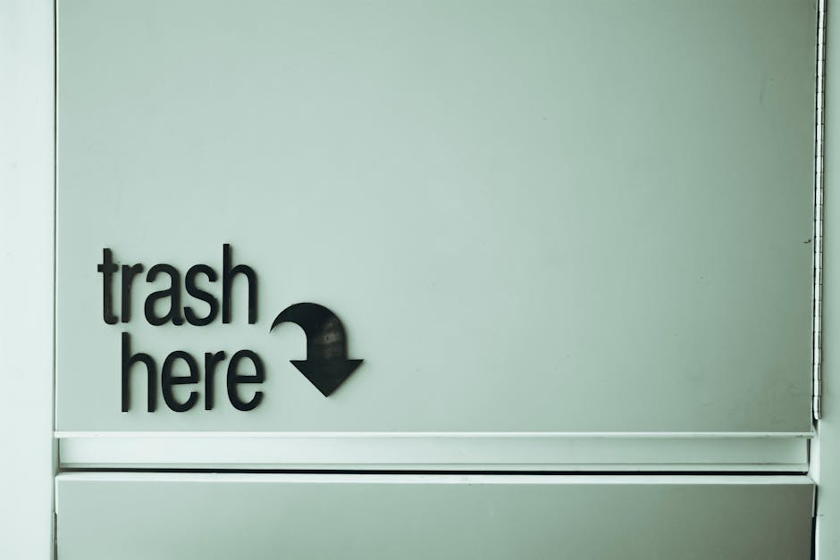A light grey wall features a black, three-dimensional sign reading 'trash here' with an downward-pointing arrow symbol. The sign is mounted on a smooth, flat surface which appears to be part of a comm